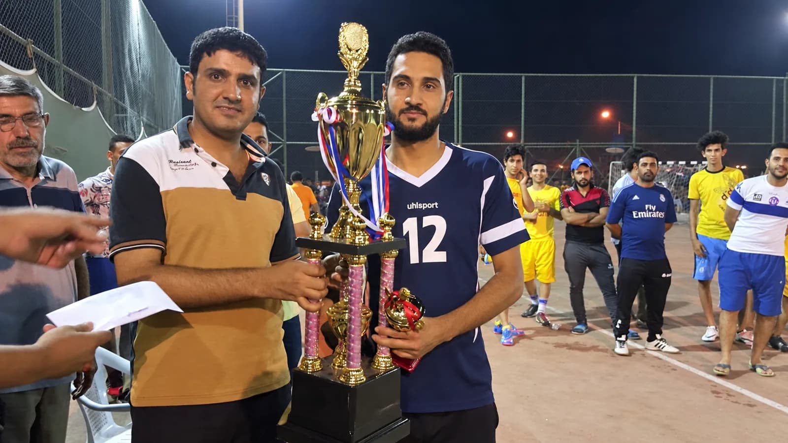 The winning captain receiving the cup from an organiser at the side of the pitch